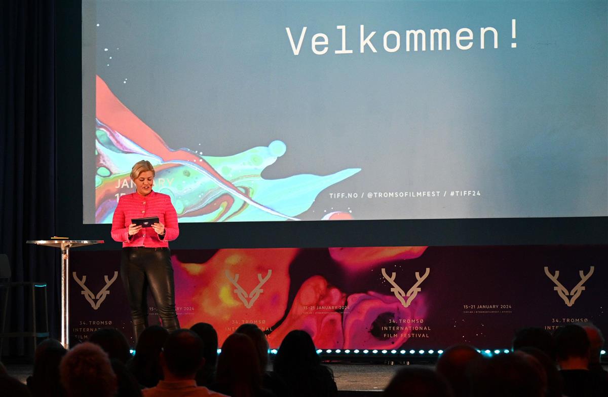En kvinne i rosa jakke står på en scene og holder tale. Fargerik bakgrunn. Foto.  - Klikk for stort bilde